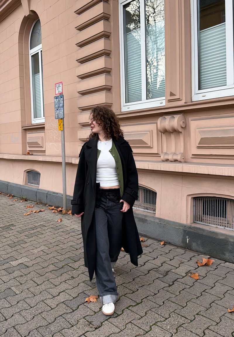 Trench-coat noir sur un haut blanc et un cardigan vert, associé à un jean large noir et des baskets blanches, marchant sur une rue pavée.