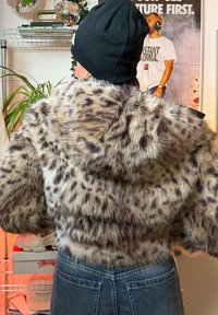Krátka bunda z umelého kožúšku hnedej farby s čiernymi leopardími škvrnami, s kapucňou a širokými rukávmi, v kombinácii s džínsami s vysokým pásom.