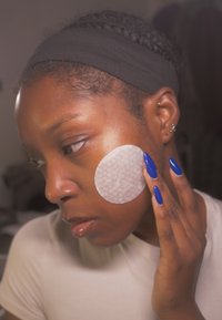 Pyöreä, kuvioitu puuvillainen kasvopyyhe iholla. Mallilla on tumma iho, pitkät siniset kynnet, vaalea toppi ja panta.