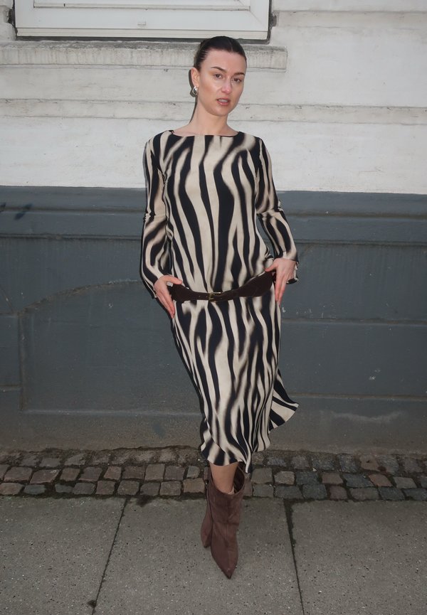 Vrouw die een lange jurk met zebra strepen draagt, een bruine riem en bruine puntlaarzen, staat op een kasseienstraat naast een grijs-witte muur.