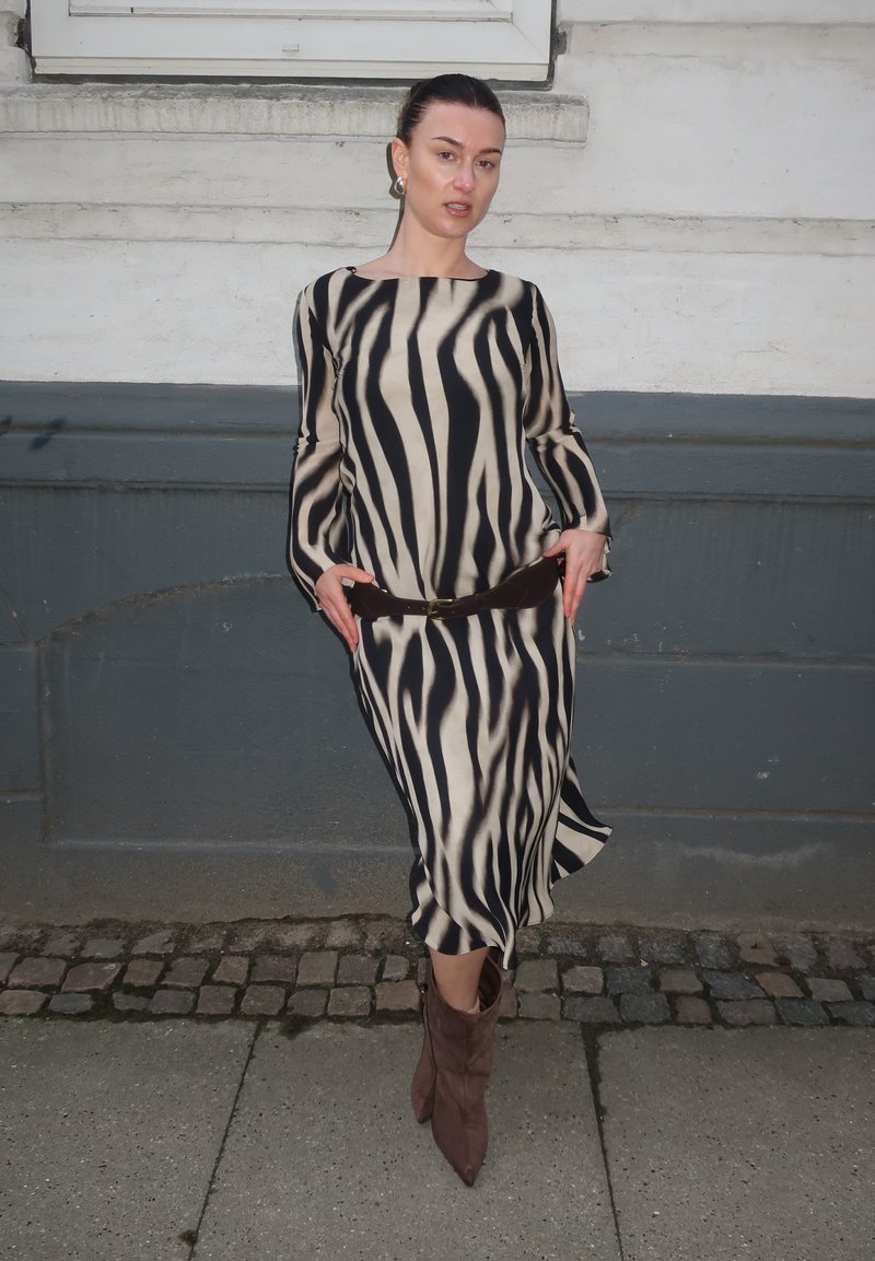 Γυναίκα με μακρύ φόρεμα με ρίγες ζέβρας, καφέ ζώνη και καφέ μυτερές μπότες στέκεται σε λιθόστρωτο πεζοδρόμιο δίπλα σε γκρι και λευκό τοίχο.