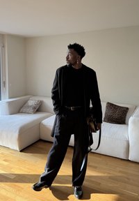 Homme en manteau, pantalon et chaussures noirs debout dans une pièce éclairée par le soleil, tenant un sac, avec un canapé d'angle blanc et des coussins à motif léopard derrière lui.