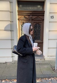 Sort uldtfrakke, grå faux pels hætte, sort håndtaske, holder en kaffekop. Står ved en brun udsmykket dør med dekorative detaljer.