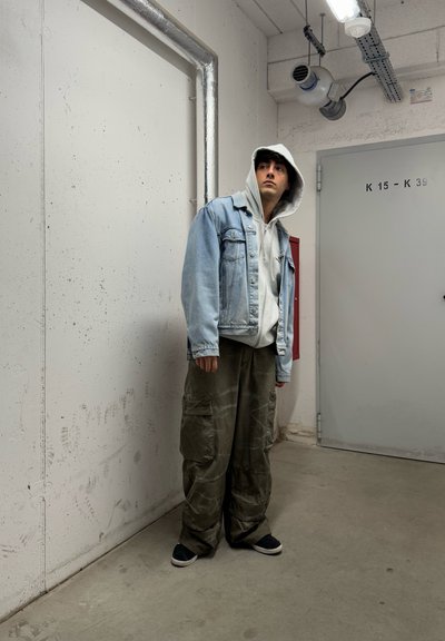 Conjunto casual que incluye una chaqueta de mezclilla azul claro, sudadera gris y pantalones cargo holgados en color oliva. Unos zapatos negros sin cordones completan el look.