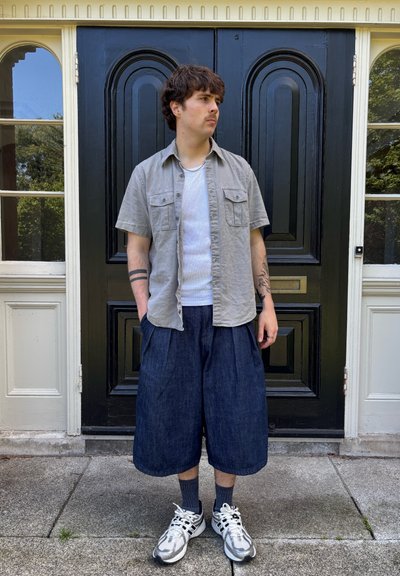 Camisa de manga corta gris claro, camiseta blanca, pantalones culotte de mezclilla azul marino, calcetines oscuros a rayas y zapatillas deportivas plateadas frente a una puerta negra.