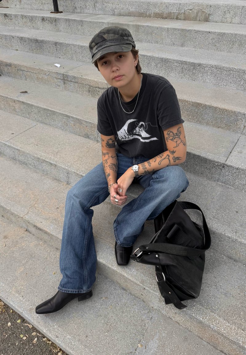 Jeune personne avec des tatouages, t-shirt graphique noir, jean, bottes noires et casquette camouflage, assise sur des marches en pierre avec un sac noir à côté.