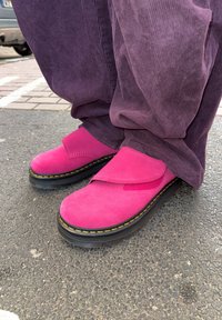Fuchsia suede slip-on shoes with a thick black sole, featuring yellow stitching and a fabric flap across the upper. Worn with purple corduroy trousers.