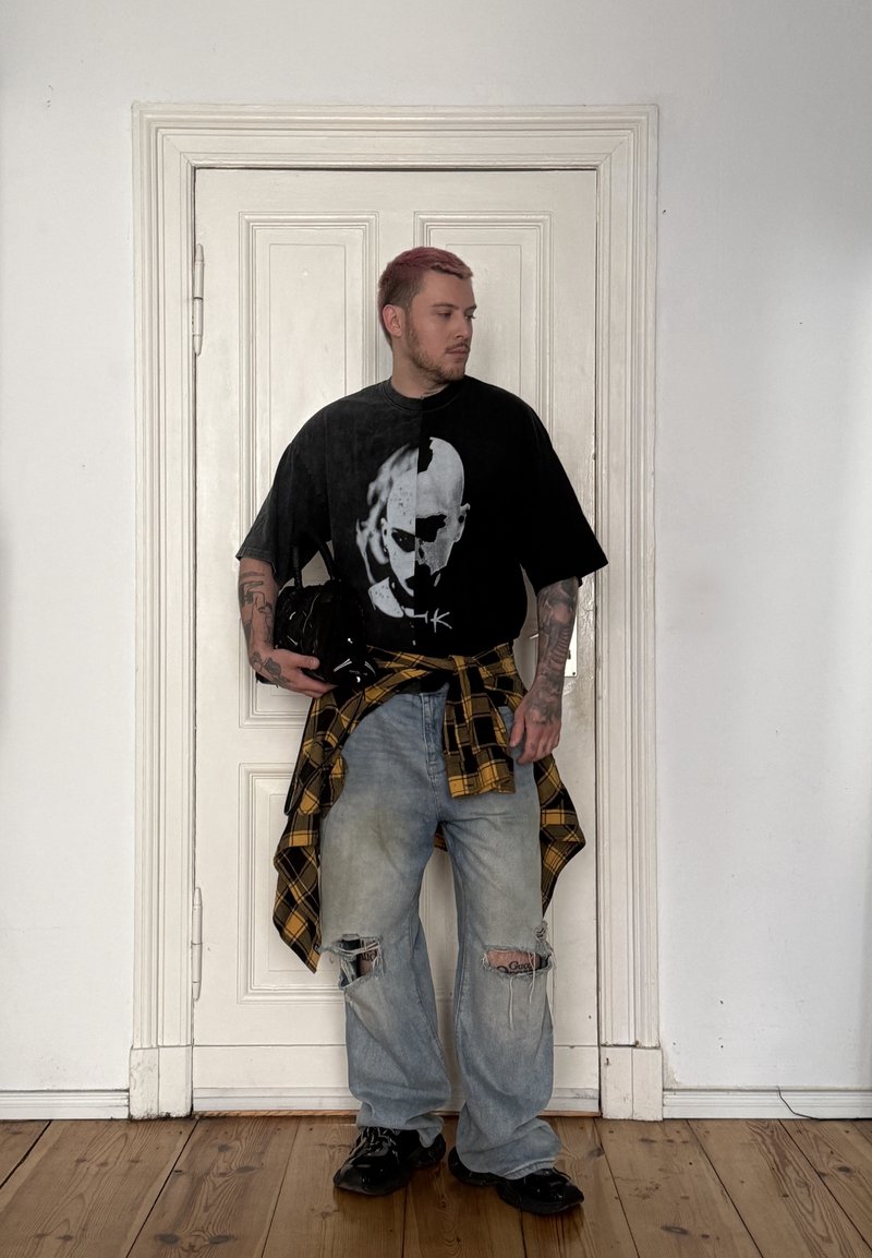 Jeune homme aux cheveux roses se tenant devant une porte blanche, portant un t-shirt noir avec un crâne, un jean déchiré et une chemise à carreaux jaune nouée autour de la taille.