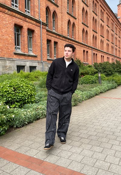 Chaqueta negra con cremallera sobre una camisa blanca, combinada con pantalones de pierna ancha a rayas blancas y negras. Zapatos negros, caminando por un camino de piedras.