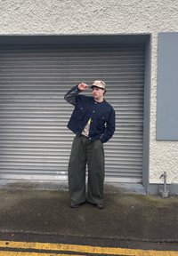 Tengerészkék farmerdzseki kockás ujjakkal, olívazöld széles szárú nadrág, és mintás sapka. Sötét cipők, szürke redőny háttér előtt pózolva.