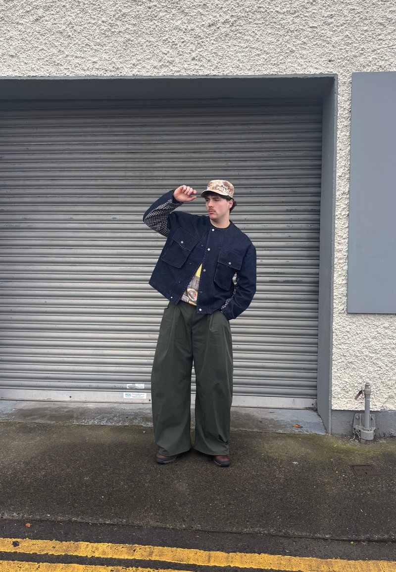 Veste en denim bleu marine avec manches à carreaux, pantalon large olive, et casquette à motifs. Chaussures foncées, posée devant un rideau métallique gris.