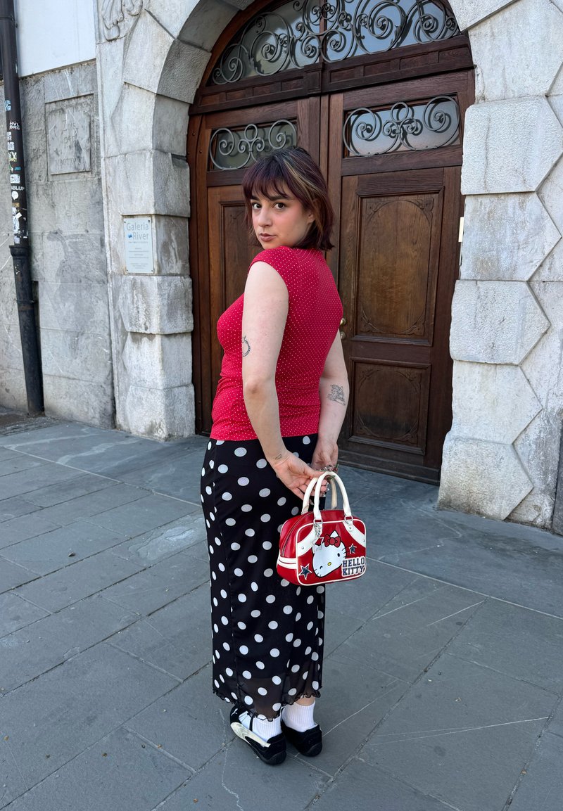 Jeune femme en haut rouge sans manches et jupe noire à pois, tenant un sac Hello Kitty, debout sur un pavé en pierre devant une porte en bois.