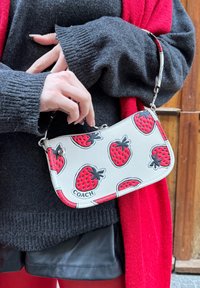 Borsa in pelle con motivo di fragole e accenti rossi, dotata di una tracolla in argento e un'etichetta con il marchio 'COACH'. Design compatto e curvato.