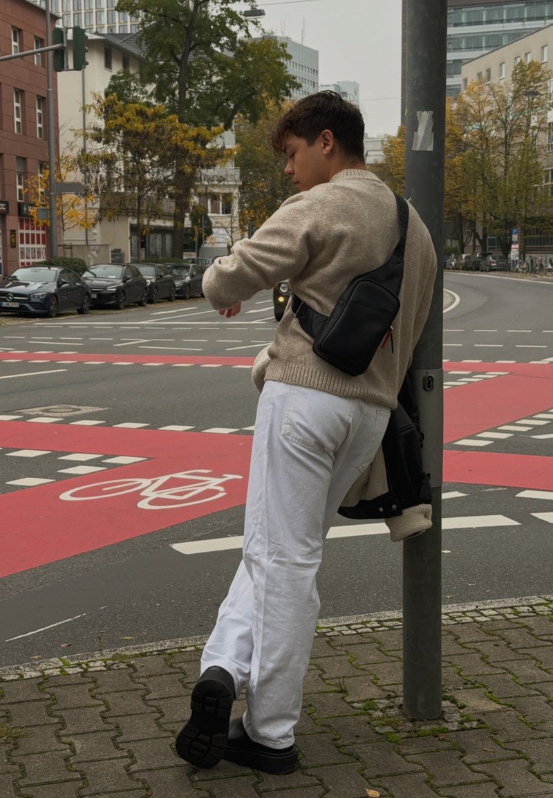 Μπεζ πλεκτή μπλούζα, λευκά παντελόνια και μαύρη τσάντα χιαστί. Στέκεται δίπλα σε μια γκρι κολώνα σε μια διασταύρωση με κόκκινες γραμμές ποδηλατοδρόμου.