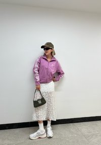 Fialová zipsová bunda s bielymi pruhmi, vrstvená biela čipkovaná sukňa, zelená taška a biele tenisky s ružovými akcentmi. Čierne slnečné okuliare a čiapka.
