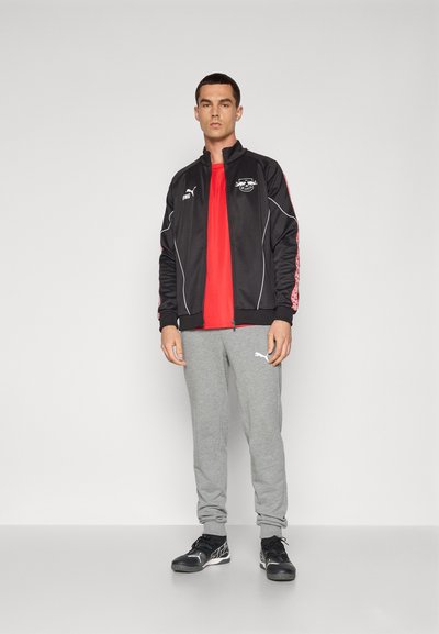 Chaqueta de track negra con detalles y logo blancos, camiseta roja brillante, pantalones de chándal grises y zapatillas deportivas negras. Diseño simple y deportivo.