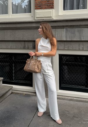 Top blanco sin mangas emparejado con pantalones anchos blancos. Sosteniendo un bolso marrón texturizado, llevando zapatillas de malla rosa, de pie al aire libre.