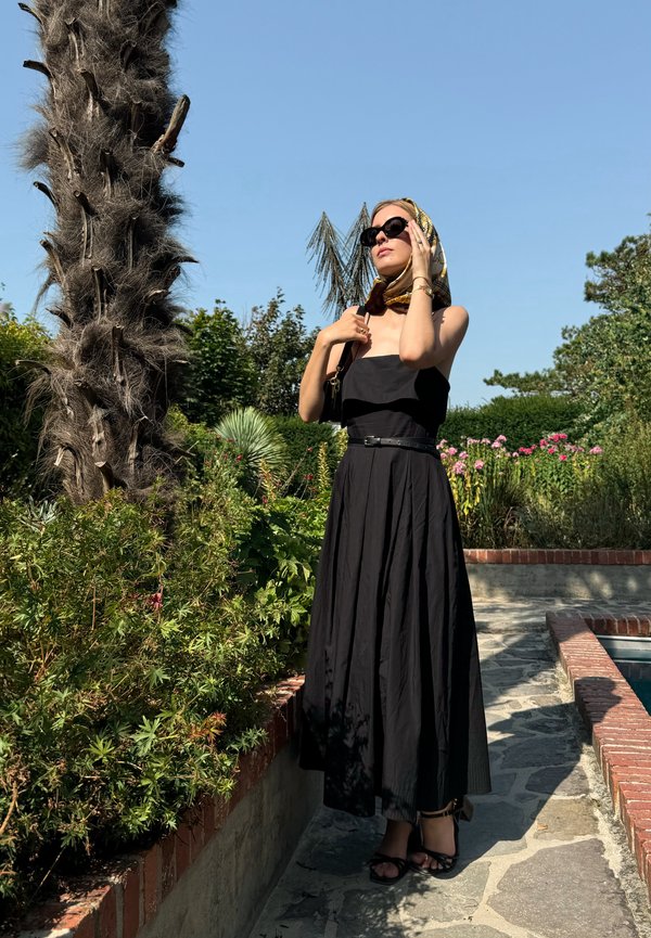 Vestido maxi negro sin mangas con un cuerpo ajustado y falda fluida; acompañado de una bufanda con estampado y gafas de sol. Se pueden ver un camino de ladrillo y plantas.