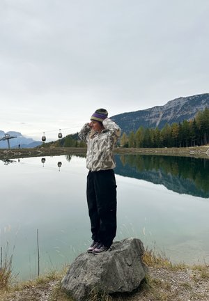 Mannequin portant une veste à motifs gris clair et beige, un pantalon noir et des chaussures violettes, debout sur un rocher près d'un lac calme et de montagnes.
