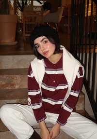 Camicia a maniche lunghe a righe marrone e bianco con colletto, sovrapposta a un maglione bianco sulle spalle, e pantaloni crema con texture. Berretto nero.