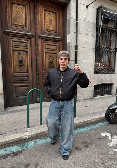 Chaqueta bomber negra con cremallera dorada, jeans de mezclilla azul sueltos y zapatos negros. El fondo presenta grandes puertas de madera y un aparcamiento para bicicletas verde.