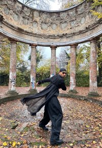 Personne en tenue noire fluide et lunettes de soleil posant de manière dynamique à l'intérieur d'un pavillon circulaire en pierre usée avec de grandes colonnes et des graffitis.