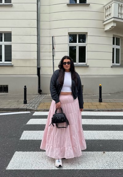 Chaqueta bomber de punto negro sobre un top corto blanco, falda larga de capas rosa, sosteniendo un pequeño bolso negro, de pie en un paso de peatones.