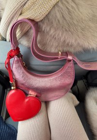 Sac à main croissant métallisé rose avec une pochette à pièces en forme de cœur rouge attachée, posé sur un pantalon en tricot crème et un fond en tissu en fourrure beige.
