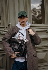 Manteau à carreaux marron porté sur un sweat à capuche gris, tenant un sac en cuir noir avec un magazine visible à l'intérieur. Une casquette vert foncé complète le look.