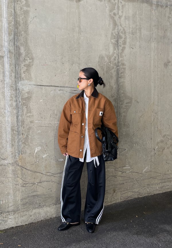 Mujer con el cabello peinado hacia atrás lleva una chaqueta marrón, camisa blanca, pantalones deportivos negros con rayas blancas, zapatos negros, gafas de sol y sostiene una bolsa negra.