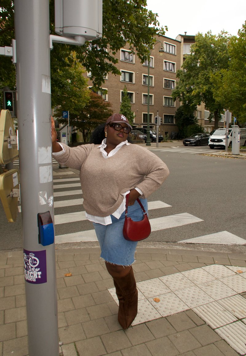 Femme portant un pull beige, une jupe en denim et des bottes brunes se tient au passage piéton en tenant un poteau, avec des arbres et des bâtiments en arrière-plan.
