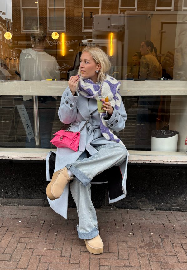 Cappotto azzurro chiaro con vestibilità oversize, jeans grigi a gamba larga, stivali beige con piattaforma. Borsa rosa, sciarpa a fantasia viola e bianca, mentre si tiene una bevanda.