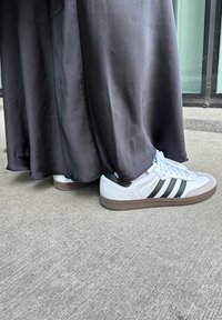 Hvide sneakers med sorte striber og en struktureret brun sål, delvist skjult af et gulvlængde sort satinplagg. Synlig betonbund.