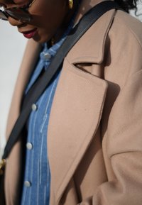 Μπεζ μάλλινο παλτό με γιακά τύπου notch πάνω από ένα μπλε ριγέ πουκάμισο με κουμπιά. Ορατός μαύρος ιμάντας ώμου, αντικατοπτρίζοντας μια casual εμφάνιση.