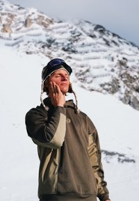Jeune homme en vêtements d'hiver et lunettes de ski se reposant les yeux fermés, touchant son visage, montagne enneigée en arrière-plan.