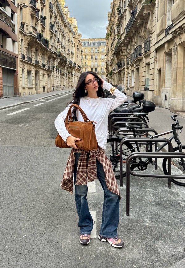 Brown suede handbag with dual handles. White long-sleeve shirt, plaid shirt tied at the waist, distressed blue jeans, and patterned sneakers.