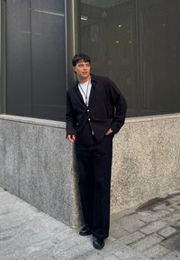 Schwarze, lässig geschnittene Hemdbluse, kombiniert mit schwarzen Hosen und Lederschuhen. Eine schlichte Silberkette setzt einen Akzent. Hintergrund mit grauer Steinmauer.