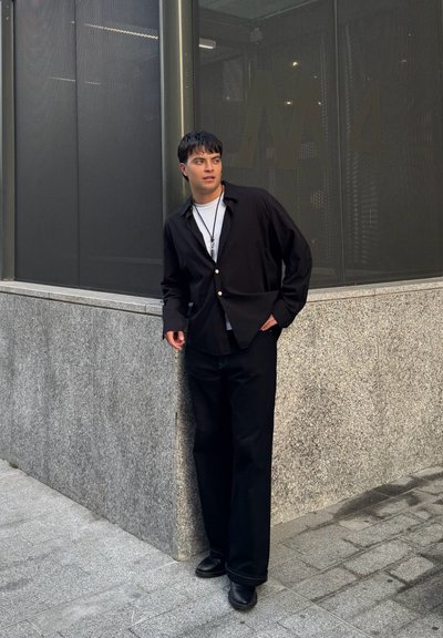 Camisa negra de botones con un corte relajado, combinada con pantalones negros y zapatos de cuero. Un collar de plata simple añade detalle. Fondo de pared de piedra gris.