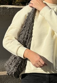 Grau strukturierte Tasche mit abgerundeten Formen und einem geschlungenen Design, die über die Schulter getragen wird von einer Person, die einen cremefarbenen Sweatshirt trägt.