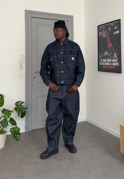 Hombre con chaqueta vaquera oscura y vaqueros holgados de pie en el interior junto a una puerta gris y un cartel motivacional de baloncesto en la pared.