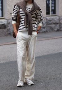White striped knit sweater with navy stripes, cream wide-leg trousers, and grey sneakers. Brown bag and sweater casually draped over the shoulders.