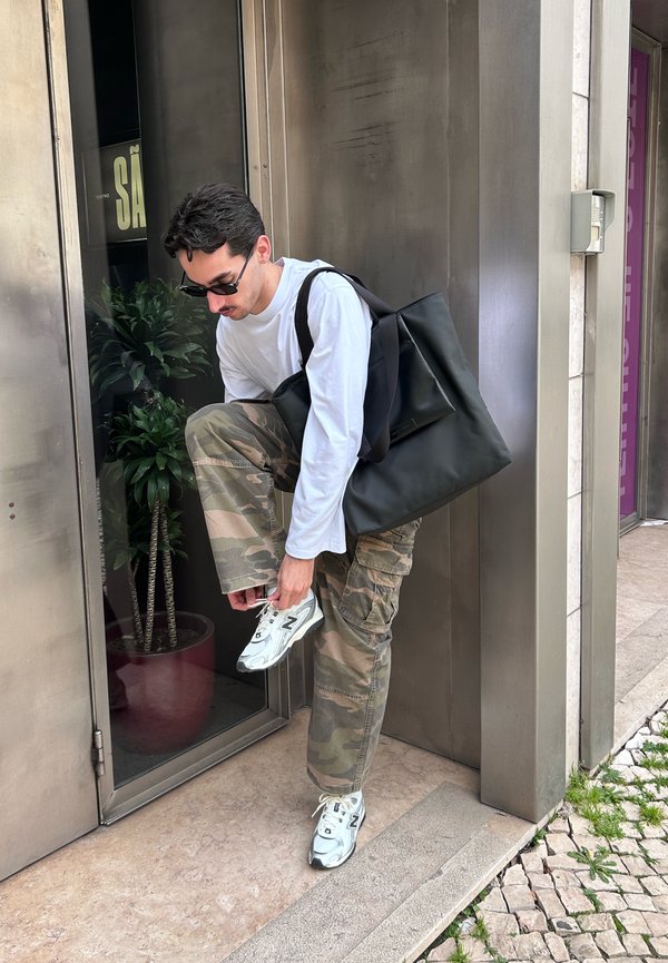 Bărbat purtând ochelari de soare și pantaloni camuflaj leagă adidaș alb lângă intrarea într-o clădire cu ușă de sticlă și o plantă în ghiveci în interior.