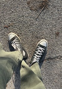 Άτομο που φοράει μαύρα ψηλά αθλητικά παπούτσια και πράσινο παντελόνι κορντούρα, στέκεται σε επιφάνεια με υφή από χαλίκι με λίγα ξερά φύλλα.