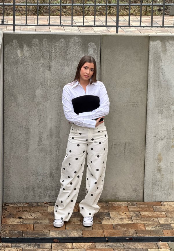 Tânără cu păr lung stă sprijinită de un perete de beton, purtând o cămașă albă, un tub negru, pantaloni albi cu buline și adidași albi.