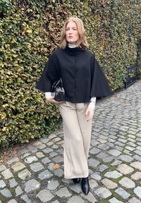 Cappa nera corta con colletto alto, pantaloni beige a gamba larga e stivali neri alla caviglia. Tiene una piccola borsa nera, ambientata su uno sfondo verdeggiante.