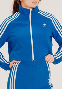 Μπλε αθλητικό μπουφάν με φερμουάρ και λευκές ρίγες, που διαθέτει ψηλό γιακά και λογότυπο Adidas. Συνδυάζεται με αντίστοιχες μπλε παντελόνες.