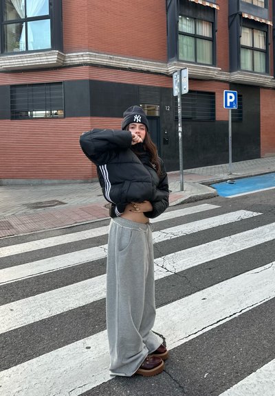 Chaqueta puffer negra con rayas blancas, pantalones de chándal grises, zapatos de plataforma marrones y un gorro negro con un logo; de pie en un paso de peatones.