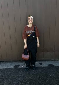 Μαύρα σαλοπέτα με καφέ γραφικό μπλουζάκι, συνδυασμένα με μωβ τσάντα. Τα αξεσουάρ περιλαμβάνουν σκουλαρίκια κρίκους και ένα κασκόλ με σχέδιο.