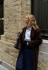 Cappotto in lana marrone oversize sopra una camicia bianca, abbinato a pantaloni in denim scuri a vita alta. Porta una borsa in pelle marrone scuro.