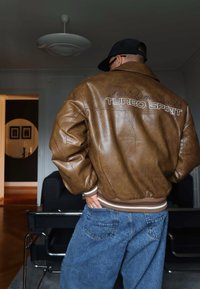 Mann iført brun skinnjakke med teksten "Turbo Spirit" på ryggen, blå jeans og svart caps, som står innendørs nær bord og stoler.